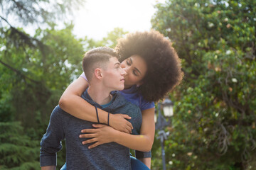 Fototapeta premium Caucasian boy carrying afro girl on his back kissing on the street with trees in the background
