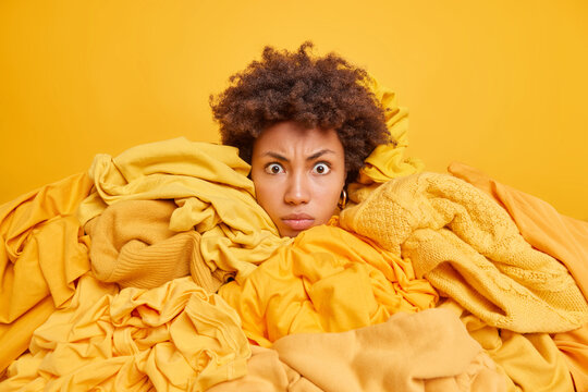 Serious curly haired Afro American woman looks shocked at camera overwhelmed with stack of yellow clothes sorts out garments accroding to color grabs apparels for washing being real shopaholic