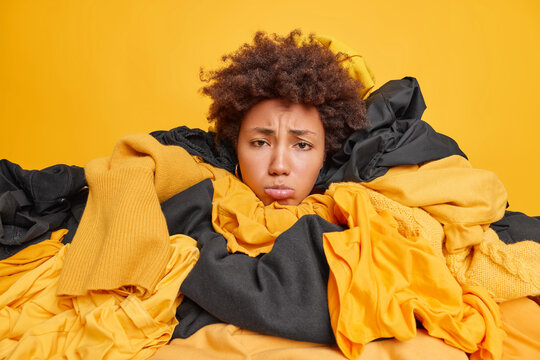 Frustrated Afro American woman covered with big pile of clothes ready for laundry collects clothing for charity or recycling has displeased face expression busy doing wardrobe cleaning. Tyding up
