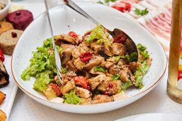 Salad with chicken and vegetables on a white plate with arugula. Lettuce leaves are mixed with chicken, vegetables and spices. salad tongs are in a plate on the salad. 