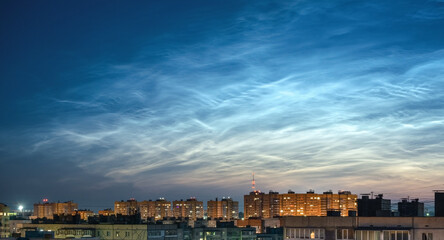 Wide panorama of the night sky and spectacular silvery clouds, silhouettes of city buildings against a background of shining clouds. Atmospheric phenomenon glowing of noctilucent clouds.