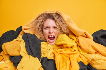 Big sale in second hand. Emotional young woman with curly hair screams loudly covered with stack of...
