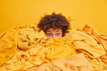 Stunned surprised African American woman covered with pile of laundry overwhelmed with clothing...