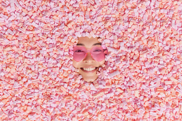 From above shot of happy cheerful woman poses around pile of pink soft sweet marshmallows enjoys eating appetizing snack wears heart shaped sunglasses. Face of smiling female model through dessert