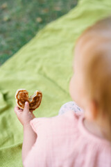 Little girl holds a bitten pancake in her hand standing on a blanket on a green lawn. Top view