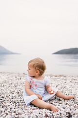 Fototapeta na wymiar Little girl in a dress sits on a pebble beach, turning her head back