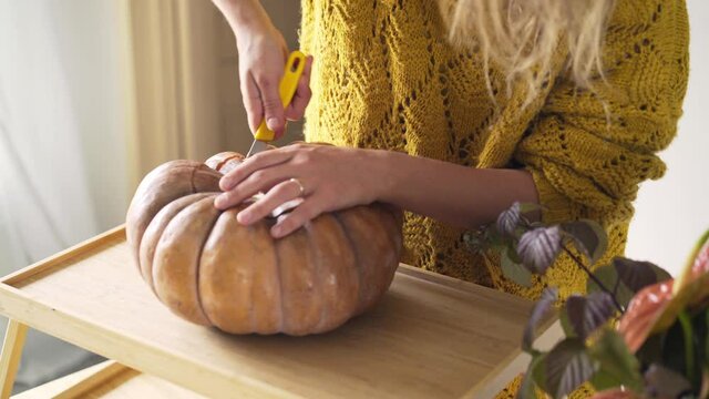 Faceless woman in yellow sweater carves scary face on large orange pumpkin with sharp knife to make traditional Halloween decor in light room. close-up shot