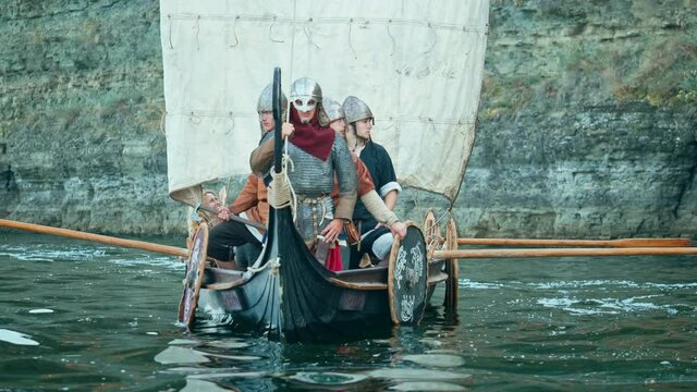 Vikings Sail on an Ancient Ship Along the River Against the Backdrop of the Rocky Coast. Men in Armor in Helmets with a Sword are Preparing to Attack. Medieval Reconstruction.