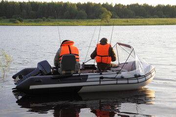 Two fishers in orange lifejackets floats on modern inflatable motor boat with 9.9 hp outboard motor that does not require registration, recreation with fishing rods on the Russian river at summer day