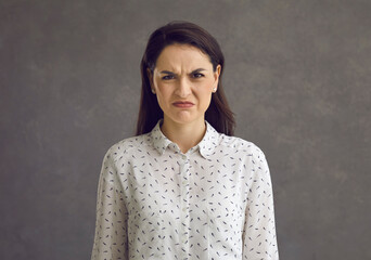 Portrait of an emotional young woman with a frowning grimace who expresses her disgust and dislike. Frustrated woman on a gray background. Concept of negative human emotions, feelings and reactions. - obrazy, fototapety, plakaty