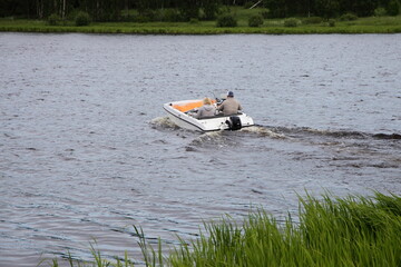 A man and a woman sail away from the grassy shore on a small white motor boat with an outboard...