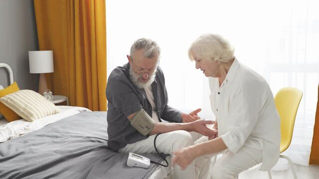 Blood pressure. Cropped gray-haired elderly woman helping her sick husband to check blood pressure using tonometer. at home in bedroom.