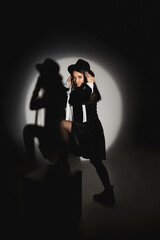 Young woman with slim body in a black trendy hat and short black dress posing in spot light at black studio
