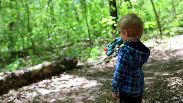 Little happy male child playing with pistol toy shooting at target in sunny green forest nature looking at camera with sunglasses. Adventure time with family. Children activity outside. Shooter.