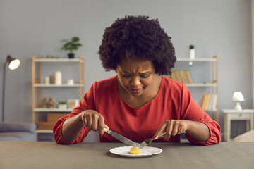 Young lady having tiny serving for lunch. Sad black woman who's sticking to strict diet eating nothing more than ridiculous portion of one little piece of apple. Nourishment and weight loss concept - obrazy, fototapety, plakaty