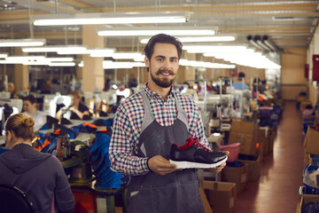 Young male shoemaker designerwith friendly smile on face wearing apron presenting new sports...