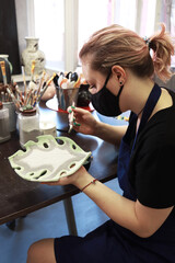 A young woman ceramist paints her clay craft. Potter at work on painting products. Creative process. Vertical photo format