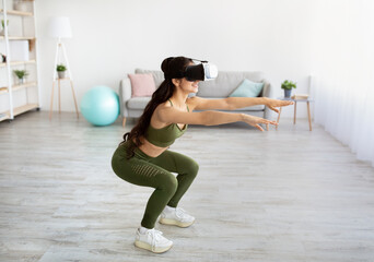 Fototapeta premium Home workout with virtual reality. Young Indian woman in VR headset doing squats at home