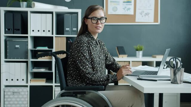 Portrait of cheerful young lady in wheelchair smiling looking at camera indoors in office. Disabled independent people and workplace concept.