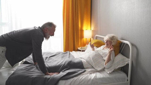 Happy Retired Man And Woman Having Fun In The Morning At Home, Gray Haired Man Amusing Wife Lying On Bed.