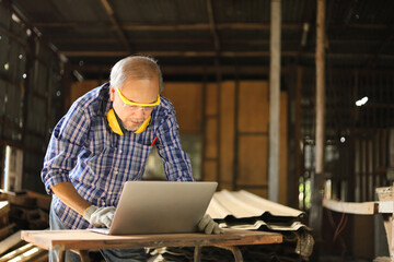 Senior Asian carpenter man is using computer laptop inside his home garage to get new modern...