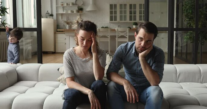 Stressed unhappy desperate frustrated young couple parents sitting on sofa, feeling tired of naughty small adopted kids running around at home, problems of raising disobedient difficult children.