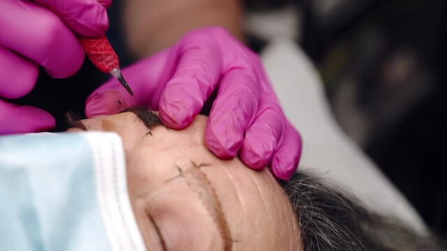 Close-up shot in slow motion of cosmetology artist using tattoo machine and dark brown colour pigment and injecting powder under the skin. Trendy microblading procedures in beauty salon, eyebrows