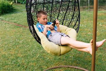 Boy in plaid shirt sitting in cocoon chair and playing with telephone. Smartphone games. Summer time