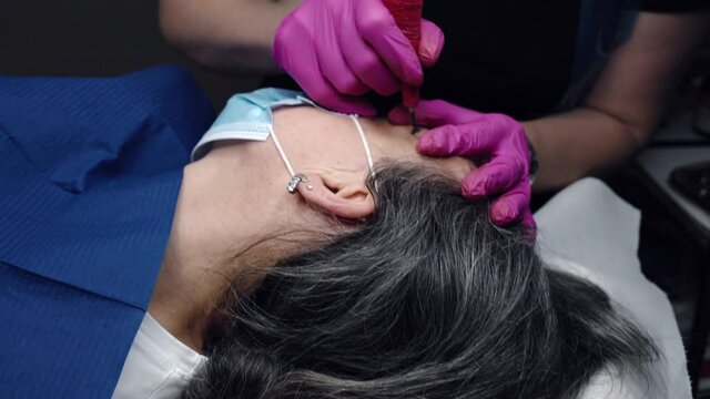 Slow motion shot of eyebrows tattoo procedure in beauty salon, cosmetology artist in protective pink gloves using tattoo machine for permanent eyebrows correction. Tattoo specialist injecting dark