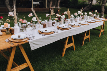 Wooden, long decorated with flowers, reeds, empty dishes, plates, forks, knives, glasses with a white tablecloth stands on the green grass in the park, forest. Wedding banquet.