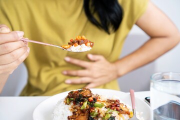 Asian woman eating spicy food and having acid reflux or heartburn hand holding a spoon with chili peppers another hand holding her stomach ache - obrazy, fototapety, plakaty