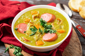 Green pea soup with carrots, potatoes, celery, sausages, pork and bacon in a white bowl on a dark wooden background. Snert is a Dutch pea soup.