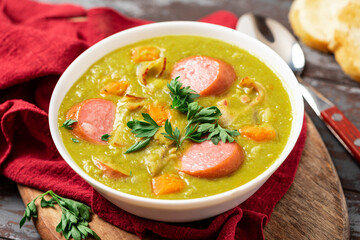 Green pea soup with carrots, potatoes, celery, sausages, pork and bacon in a white bowl on a dark...