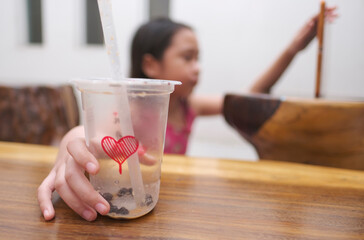 The bubble milk tea plastic cup with love symbol almost empty. A girl is holding a plastic cup drink straw with her right hand, left hand is holding a tablet as a bokeh background.