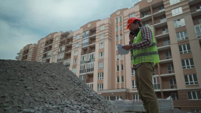 Satisfied foreman in helmet and vest at construction site talking on phone with rubble contractor and looks at waybills. Builder will check arrival building material, check shortage and call by phone.