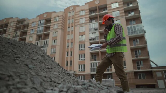 Inspecting builder speaks angrily on phone with subordinate at construction site checks amount of crushed stone for the construction of road in residential area according to incoming invoices.