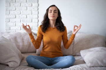 Cute concentrated young smiling beautiful brunette woman is meditating in lotus pose on the couch...