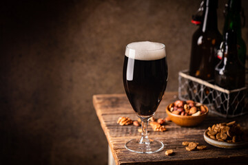 Glass of dark beer with foam head on dark wooden background with empty bottles and beer snacks