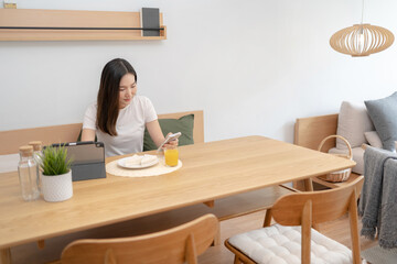 Happy asian woman using mobile phone during breakfast or eating lunch while working from home.