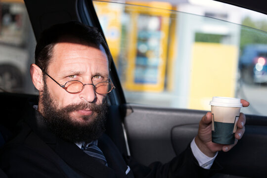 Brunette Bearded Man In Glasses, White Shirt And Business Suit Sitting At His Car Driving And Drinking Coffee To Go On A Sunny Day. Shadow And Light, Sunlight Over The Man's Face. Coffee And Drive