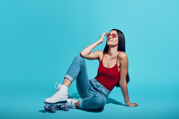 Attractive young woman wearing roller skates and smiling while sitting against blue background