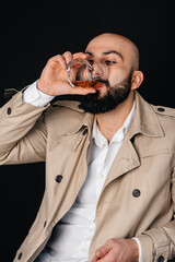 A young man sits on a black background and drinks an alcoholic drink.