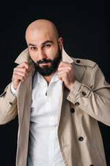 A young bearded man stands and adjusts his raincoat against a black background.