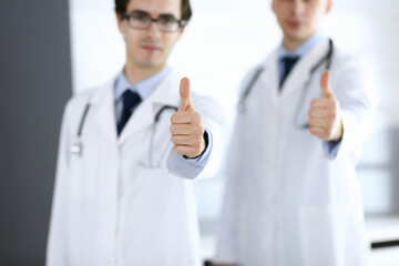 Two male doctors standing as a team and showing thumbs up as a symbol of the best service for patients in the clinic. Medicine and health care