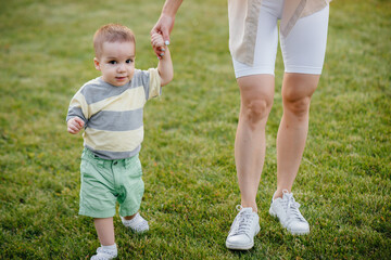 A young cute mother helps and teaches her little son to take his first steps during sunset in the park on the grass.