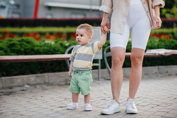 A young cute mother helps and teaches her little son to take his first steps during sunset in the park on the grass.