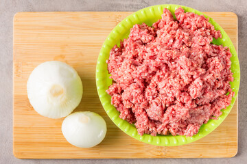 Chopped minced meat in a green plastic plate and onions on a cutting wooden board. View from above.