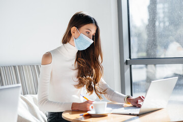 A young girl is sitting in a cafe wearing a mask and working at a computer.