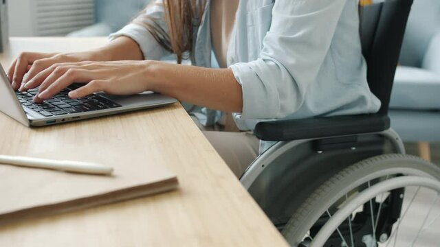 Slow motion of female student learning online using laptop typing sitting in wheelchair in apartment busy with distant education. Technology and disability concept.
