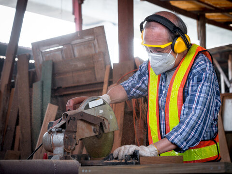 Elderly carpenter craftsman wearing safety gear working with hardwood using a miter saw to cut a piece of wood in the workshop. Manufacture of wood products.. - Powered by Adobe